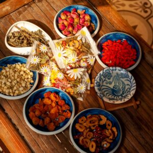 Assorted dried herbs, flowers, and fruits in colorful ceramic bowls, including rosebuds, jasmine, goji berries, and dried fruit slices, prepared for making soothing sleep blend tea
